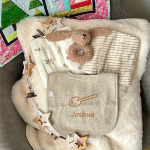 Könnte beinhalten: Ein beiger Baby-Lätzchen mit dem Namen "Joshua" bestickt, einer Löffelgrafik und einer Holzstern-Girlande. Das Lätzchen liegt auf einer weißen Kunstfelldecke mit einem braunen und weißen gestreiften Baby-Spucktuch und einem braunen Hasen-Rassel.