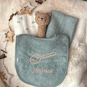Könnte beinhalten: Ein hellblauer Baby-Lätzchen mit einem Löffel und dem Namen Joshua in rosa gestickt. Das Lätzchen liegt auf einer weißen, flauschigen Oberfläche mit einem weißen und grauen gemusterten Baby-Spucktuch, einem Plüsch-Teddybär und einem weißen und grauen gemusterten Baby-Spucktuch mit Hasen.