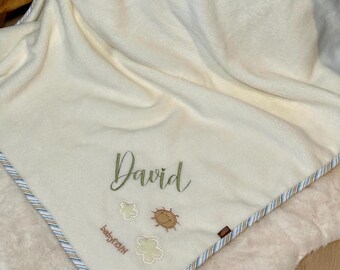 Op de afbeelding: Een crèmekleurige fleecedeken met de geborduurde naam David in groen schrift. Onder de naam staan twee kleine witte wolkenomtrekken en een bruin zonicoon. De deken heeft een decoratieve rand met dunne blauwe en bruine strepen.