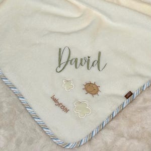 Può includere: Coperta per bambini color crema con il nome "David" ricamato in verde. La coperta presenta ricami di sole e fiori e un bordo a righe blu, marrone e bianco. La coperta ha una consistenza morbida e soffice.