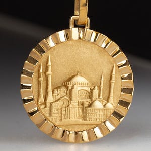 May include: Gold pendant necklace. The circular pendant features a detailed relief of a building with minarets and domes, possibly a mosque. The pendant has a textured border and hangs from a rope chain.