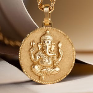 May include: A gold-colored pendant necklace featuring a detailed depiction of the Hindu deity Ganesha. The pendant is round with a beaded border and hangs from a rope chain. The image is set against a soft, neutral background.