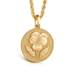 May include: A gold-toned necklace featuring a circular pendant with a raised pansy flower design. The pendant has a beaded edge and is suspended from a twisted rope chain. The entire piece has a matte, metallic finish with consistent gold coloring.