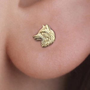 Puede incluir: Un pendiente de botón dorado con forma de cabeza de lobo se muestra en una oreja. El pendiente es pequeño y detallado, con textura de pelaje visible. Es una joya con temática de la naturaleza.
