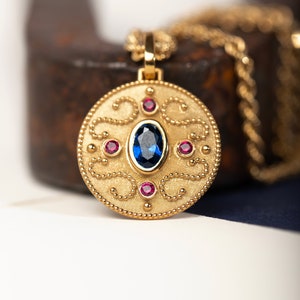 May include: A gold pendant necklace with a round, ornate design. The pendant features a blue gemstone surrounded by smaller red gemstones.