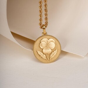 May include: A gold-toned necklace featuring a circular pendant with a raised pansy flower design. The pendant has a beaded edge and is suspended from a twisted rope chain. The metal surface has a matte finish, highlighting the floral detail.