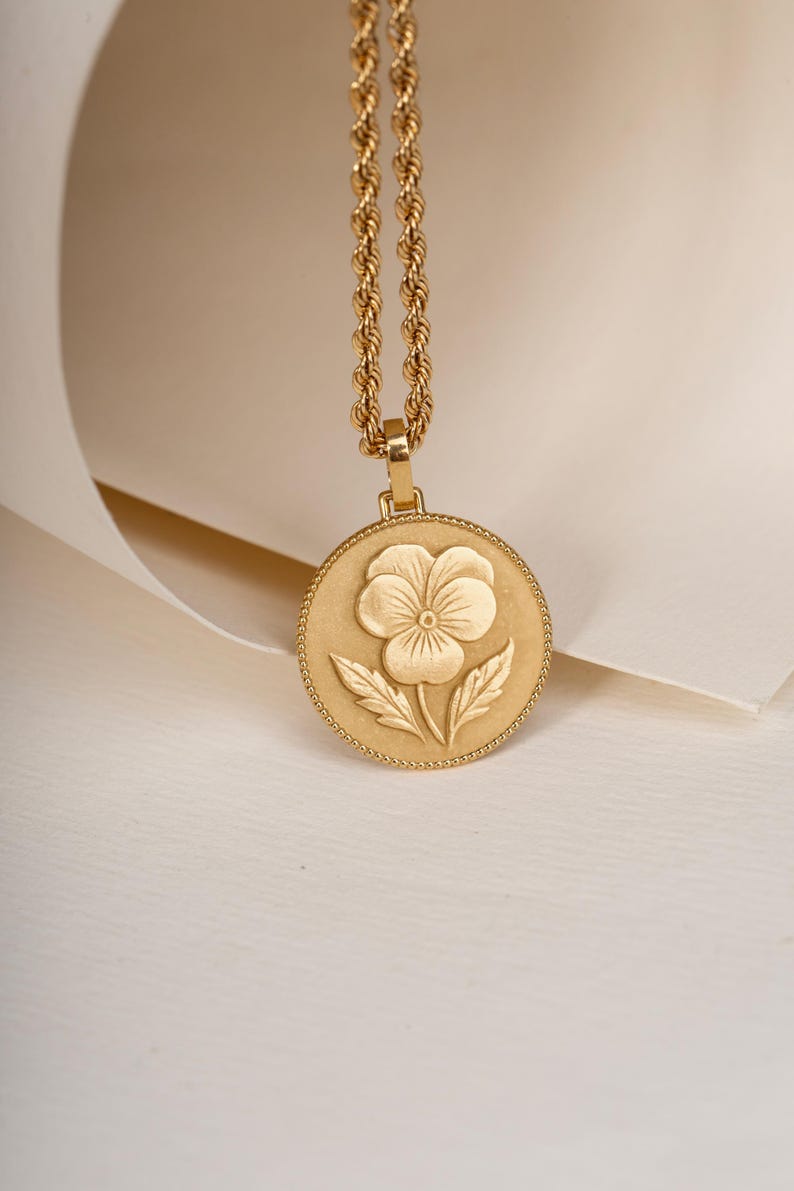 May include: A gold-toned necklace featuring a circular pendant with a raised pansy flower design. The pendant has a beaded edge and is suspended from a twisted rope chain. The metal surface has a matte finish, highlighting the floral detail.
