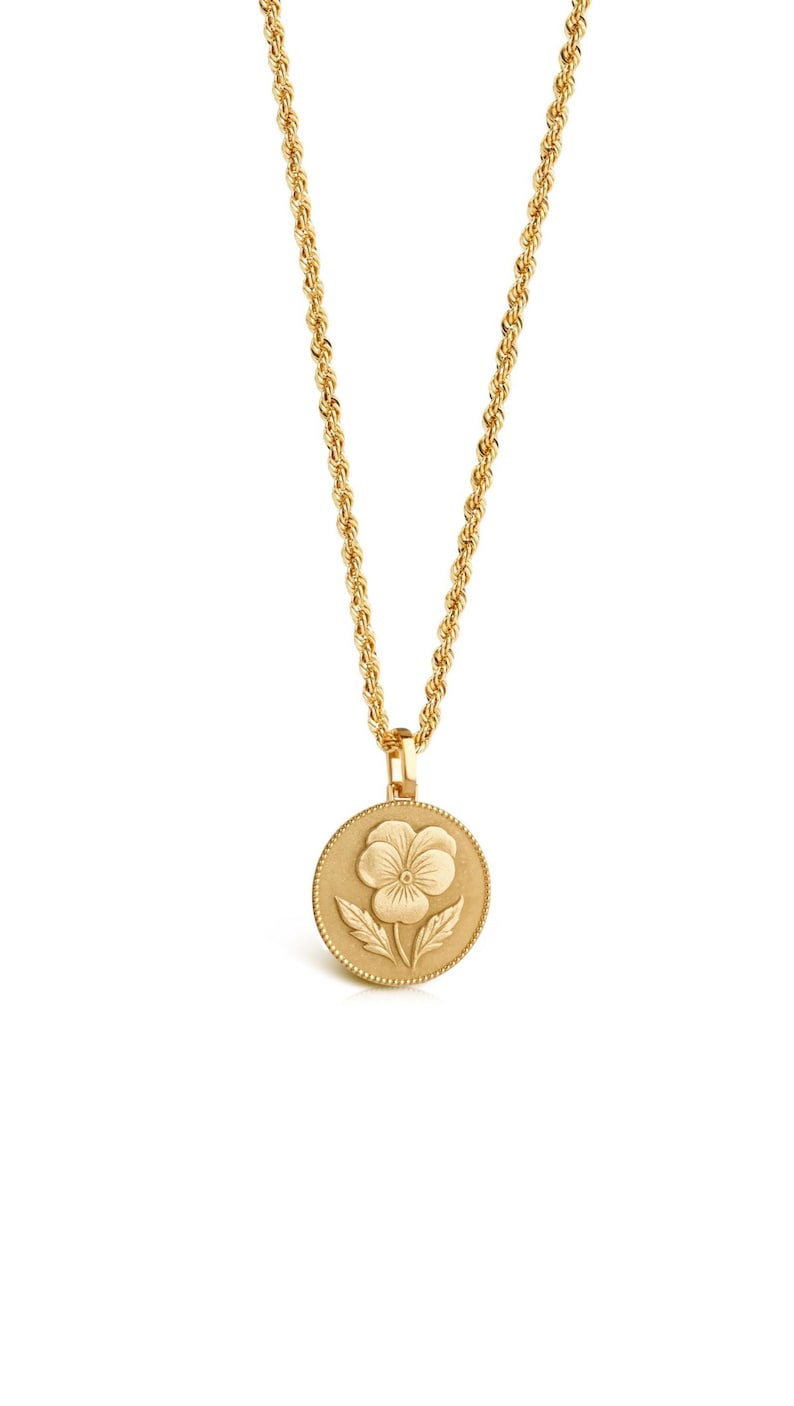 May include: A gold-toned necklace featuring a circular pendant with a raised pansy flower design. The pendant has a beaded edge and is suspended from a twisted rope chain. The entire piece has a matte, metallic finish with consistent gold coloring.