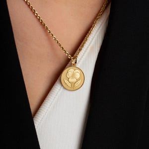 May include: A gold-toned circular pendant necklace featuring an embossed pansy flower design. The pendant hangs from a delicate twisted rope chain. The coin-shaped charm has a textured, beaded border surrounding the central floral motif.