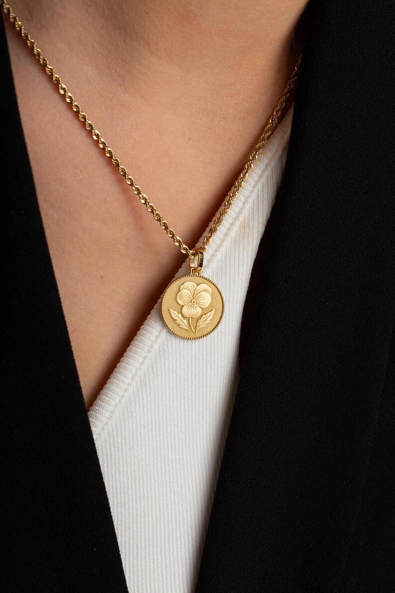 May include: A gold-toned circular pendant necklace featuring an embossed pansy flower design. The pendant hangs from a delicate twisted rope chain. The coin-shaped charm has a textured, beaded border surrounding the central floral motif.