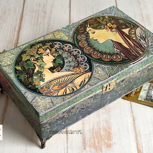 May include: A green wooden box with a hinged lid and decorative brass accents. The box is decorated with two images of women in Art Nouveau style, surrounded by floral and geometric patterns. The box is sitting on a wooden surface.