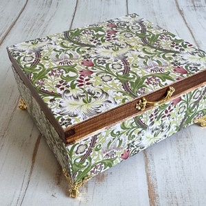 May include: A decorative wooden box with a floral pattern in green, white, and burgundy. The box has a wooden frame, gold-colored metal feet, and a handle. The box is closed and sits on a light-colored wooden surface.