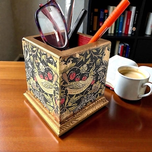 Puede incluir: Un organizador de escritorio cuadrado con un borde dorado y un patrón decorativo con pájaros, fresas y follaje sobre un fondo negro. El organizador contiene bolígrafos, gafas y un bolígrafo. Una taza de café y un libro están en el fondo.