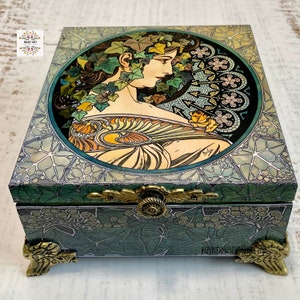 Peut inclure: Une boîte en bois verte avec des accents dorés et un couvercle à charnières. Le couvercle présente un motif circulaire avec le portrait d'une femme entouré de lierre et un motif floral. La boîte a des pieds dorés et un loquet en laiton.