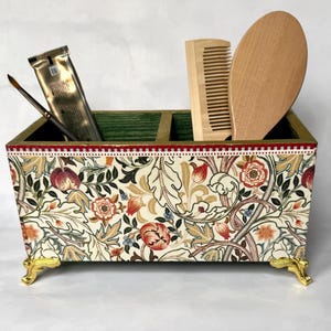 Könnte beinhalten: Eine dekorative Holzkiste mit floralem Muster und goldenen Füßen. Die Kiste hat zwei Fächer und ist gefüllt mit einem Holzlöffel, einem Kamm und einer Tube Haarprodukt.