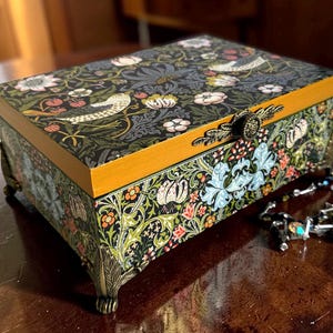 May include: A rectangular jewellery box with a floral and bird pattern on a black background. The box has a gold-coloured trim and ornate metal hardware. The box is resting on four small feet.