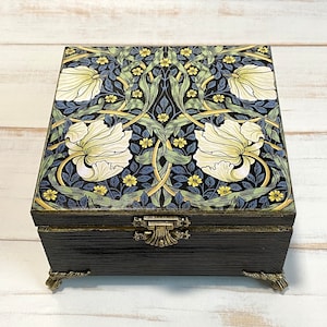Könnte beinhalten: Eine schwarze Holzkiste mit einem floralen Design auf dem Deckel. Das Design zeigt weiße Blumen, grüne Blätter und gelbe Akzente. Die Kiste hat goldene Metallfüße und einen goldenen Metallverschluss.