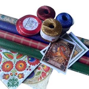 May include: A collection of craft supplies including rolls of patterned wrapping paper, spools of metallic thread in red, gold, and blue, and decorative cards featuring an Art Nouveau design. A red ribbon with white text is also visible.