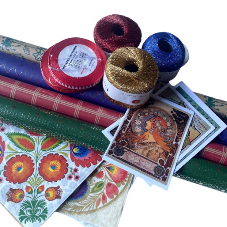 May include: A collection of craft supplies including rolls of patterned wrapping paper, spools of metallic thread in red, gold, and blue, and decorative cards featuring an Art Nouveau design. A red ribbon with white text is also visible.