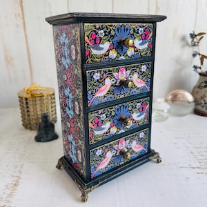 Puede incluir: Un pequeño y decorativo mueble de cajones con cuatro cajones. El mueble está pintado de color verde azulado oscuro con un estampado floral y de pájaros en rosa, azul y blanco. Los tiradores de los cajones son de color latón y el mueble tiene patas decorativas.