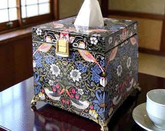 May include: A square tissue box holder featuring a dark black background with a colorful botanical pattern of birds, strawberries, and blue flowers. The box has a gold-toned metal latch on the front and stands on four ornate gold-toned metal feet.
