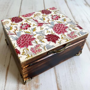 May include: A wooden box with a hinged lid and a floral pattern on the top. The box has a brass handle and decorative feet.