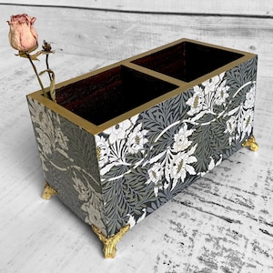 May include: A wooden desk tidy with two sections, decorated with a floral pattern in white, grey, and green. It has gold edging and feet. A dried rose is placed in one of the compartments. The organizer is approximately 20 cm long.