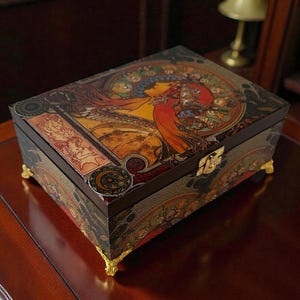 May include: A rectangular wooden jewellery box with a decorative lid featuring an Art Nouveau-style illustration of a woman with flowing red hair. The box has gold-coloured feet and a clasp. The sides are adorned with floral patterns.