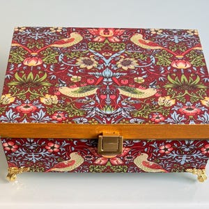 Op de afbeelding: Een rechthoekige sieradendoos met een levendig bloemen- en vogelpatroon in rood, groen, geel en blauw. De doos heeft een houten frame, goudkleurige hardware en sierlijke gouden pootjes. Een klein bloementakje en een steen liggen ernaast.