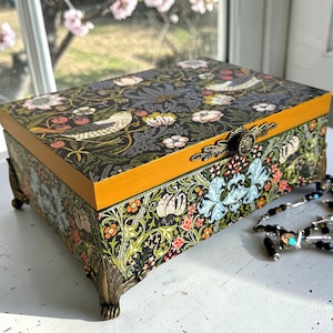 Puede incluir: Una caja de joyería rectangular de madera con un estampado floral y de pájaros sobre un fondo oscuro. La caja tiene detalles y patas de metal color latón. La tapa y el borde son de color amarillo-naranja cálido. Un collar de cuentas descansa junto a la caja.