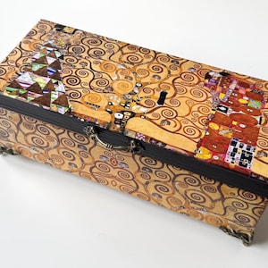 Puede incluir: Una caja de madera rectangular con tapa abatible y asa de metal. La caja está decorada con una impresión colorida de la pintura "El Árbol de la Vida" de Gustav Klimt.