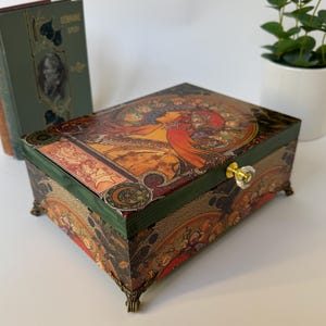 May include: A decorative wooden box with a colourful Art Nouveau design. The box features a green frame, brass feet, and a crystal knob. The lid is adorned with a vibrant illustration of a woman with flowing hair and floral elements.