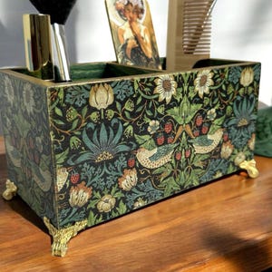 May include: A wooden organiser with four compartments, featuring a floral and bird design in shades of green, blue, and red. The organiser has gold-coloured feet and is sitting on a wooden surface.