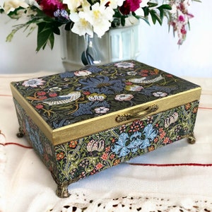 Op de afbeelding: Een decoratieve houten doos met een bloemenpatroon en gouden afwerking. De doos heeft een scharnierend deksel met een messing handvat en staat op vier gouden pootjes.