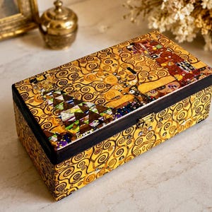 Könnte beinhalten: Eine rechteckige Holzschatulle mit goldfarbener Oberfläche, verziert mit komplexen schwarzen Spiralmustern und einer bunten Mosaik-Einlage auf dem Deckel. Die Schatulle hat einen dunkelbraunen Rand und einen kleinen Metallverschluss an der Vorderseite.
