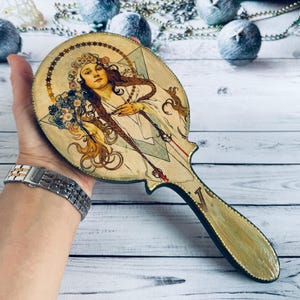 Pode incluir: Espelho de mão decorativo com uma ilustração em estilo Art Nouveau de uma mulher com cabelo comprido e uma coroa de flores. A pega é dourada e o espelho tem uma moldura de madeira com um design detalhado.