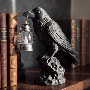 Peut inclure: Une figurine de corbeau noir mat perchée sur une base en roche texturée. Le corbeau tient dans son bec une petite lanterne argentée. La lanterne présente un globe en verre transparent et une lumière blanche en forme de bougie à l'intérieur.