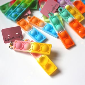 Könnte beinhalten: Regenbogen-Pop-It-Ohrringe mit silbernen Haken. Die Ohrringe sind aus Silikon und haben einen Farbverlauf. Die Ohrringe werden auf einer weißen Oberfläche präsentiert.