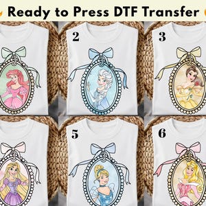 以下が含まれることがあります： ディズニーのプリンセスが描かれたDTF転写の白いTシャツ。各シャツには、リボンとリボンで飾られた楕円形のフレームの中に、異なるプリンセスが描かれています。プリンセスには、アリエル、エルサ、ベル、ラプンツェル、シンデレラ、オーロラが含まれます。「Ready to Press DTF Transfer」の文字が上部にあります。