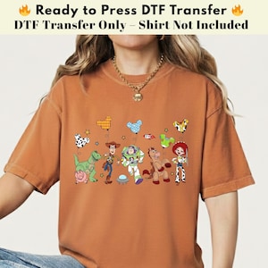 Pode incluir: Uma t-shirt de cor laranja queimado com uma impress&atilde;o gr&aacute;fica das personagens de Toy Story, incluindo Woody, Buzz Lightyear, Jessie, Rex, Bullseye e Hamm, acompanhadas por bal&otilde;es coloridos em forma de orelhas de rato e estrelas.
