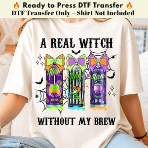 May include: A cream-colored t-shirt featuring a graphic print with the text A REAL WITCH WITHOUT MY BREW. The design includes three energy drink cans topped with colorful bows, surrounded by spiderwebs, bats, and small star icons.