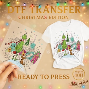 May include: White t-shirt with a Grinch-themed Christmas design. The design features the Grinch, a dog, and a whimsical village scene. The image also shows a DTF transfer sheet with the same design, and the text "READY TO PRESS".
