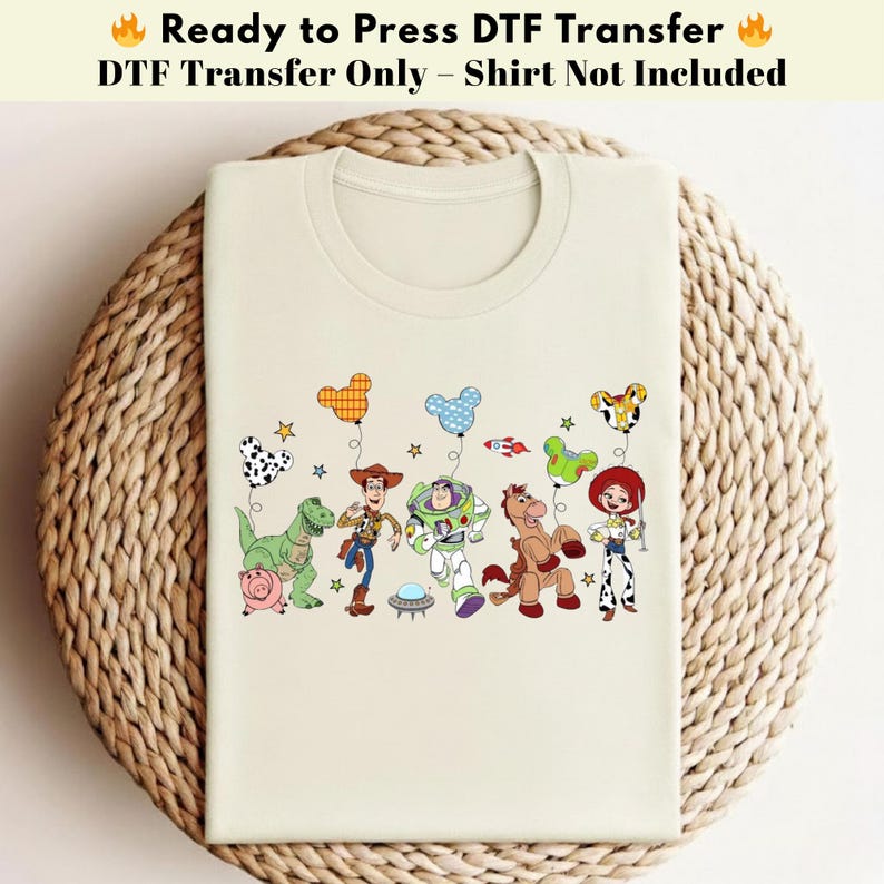 Pode incluir: Uma t-shirt de cor creme com uma impress&atilde;o gr&aacute;fica das personagens de Toy Story, incluindo Woody, Buzz Lightyear, Jessie, Rex, Bullseye e Hamm. As personagens seguram bal&otilde;es coloridos em forma de orelhas de rato e pequenas estrelas.