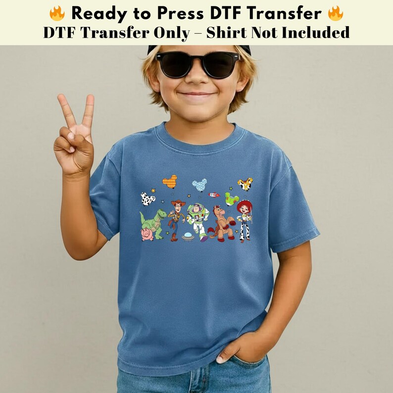 Pode incluir: Uma t-shirt azul com uma impress&atilde;o gr&aacute;fica de v&aacute;rias personagens animadas, incluindo um cowboy, um ranger espacial, uma cowgirl, um dinossauro e um porco, acompanhados por pequenos &iacute;cones de bal&otilde;es coloridos. O texto diz Ready to Press DTF Transfer.