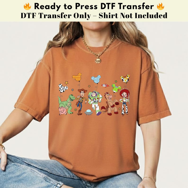 Pode incluir: Uma t-shirt de cor laranja queimado com uma impress&atilde;o gr&aacute;fica das personagens de Toy Story, incluindo Woody, Buzz Lightyear, Jessie, Rex, Bullseye e Hamm, acompanhadas por bal&otilde;es coloridos em forma de orelhas de rato e estrelas.