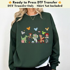 Pode incluir: Uma camisola de manga comprida verde-escura com uma impress&atilde;o gr&aacute;fica das personagens de Toy Story, incluindo Woody, Buzz Lightyear, Jessie, Rex, Bullseye e Hamm, acompanhados por bal&otilde;es coloridos em forma de Mickey e um pequeno foguet&atilde;o.