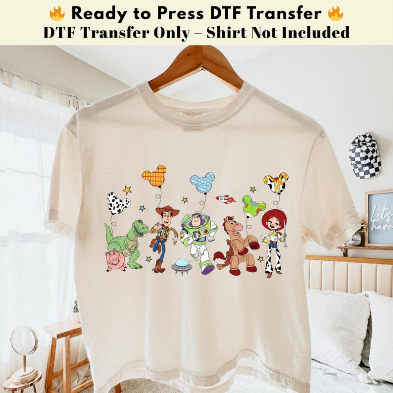 Pode incluir: Uma t-shirt de manga curta na cor creme com uma impress&atilde;o gr&aacute;fica das personagens de Toy Story, incluindo Woody, Buzz Lightyear, Jessie, Rex, Bullseye e Hamm, cada uma segurando um bal&atilde;o colorido em forma de Mickey Mouse com estrelas e um foguet&atilde;o.