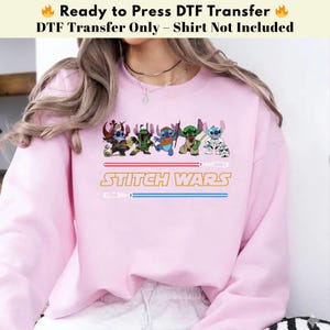Puede incluir: Una sudadera rosa claro con un dise&ntilde;o de "Stitch Wars". El dise&ntilde;o presenta personajes de dibujos animados disfrazados de personajes de Star Wars, con el texto "STITCH WARS" en dorado y l&iacute;neas azules estilo sable l&aacute;ser.