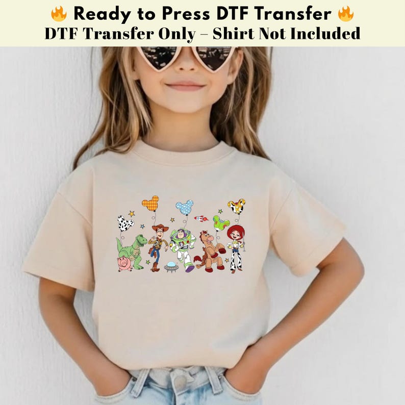 Pode incluir: Uma t-shirt bege com uma impress&atilde;o gr&aacute;fica de v&aacute;rias personagens animadas, incluindo um cowboy, um ranger espacial, uma cowgirl, um dinossauro verde, um porco e um cavalo, cada um segurando um bal&atilde;o em forma de cabe&ccedil;a de rato.