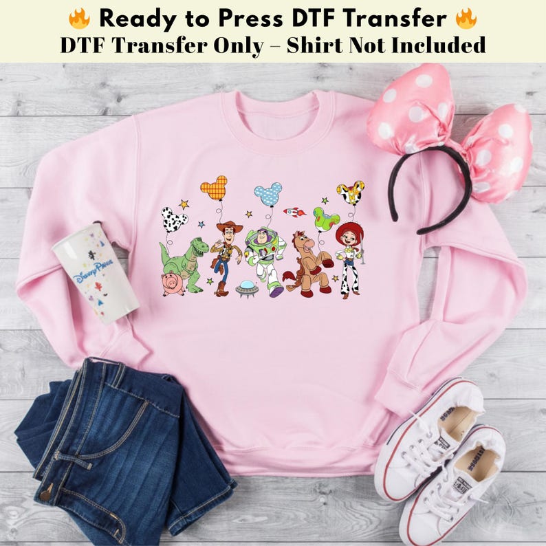 Pode incluir: Uma camisola rosa claro com uma impress&atilde;o gr&aacute;fica de personagens de Toy Story, incluindo Woody, Buzz Lightyear, Jessie, Rex, Bullseye e Hamm segurando bal&otilde;es em forma de orelhas do Mickey, com um copo branco, bandolete com la&ccedil;o e sapatilhas.